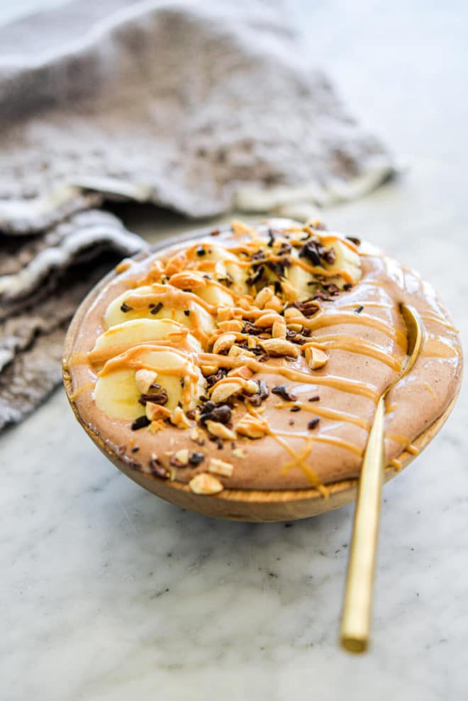 Chocolate smoothie with gold spoon in bowl topped with cocoa nibs, chopped peanuts, sliced banana, and a drizzle of peanut butter