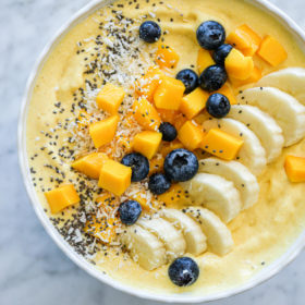 Mango smoothie in a bowl topped with sliced banana, chopped mango, blueberries, chia seeds, and coconut flakes
