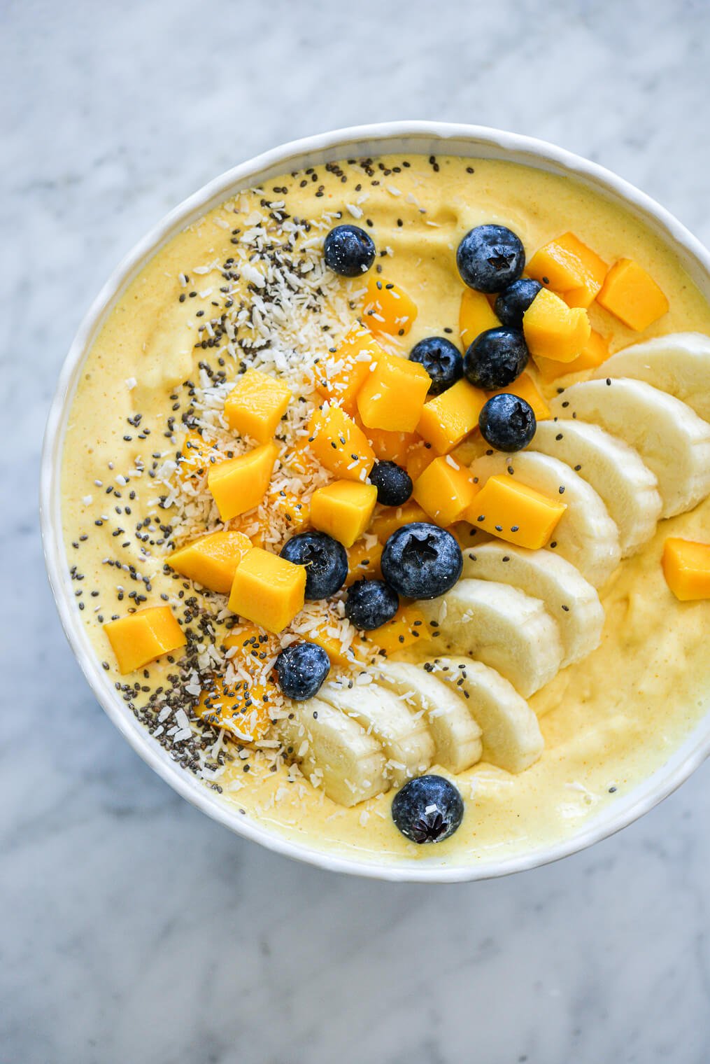 Mango smoothie in a bowl topped with sliced banana, chopped mango, blueberries, chia seeds, and coconut flakes