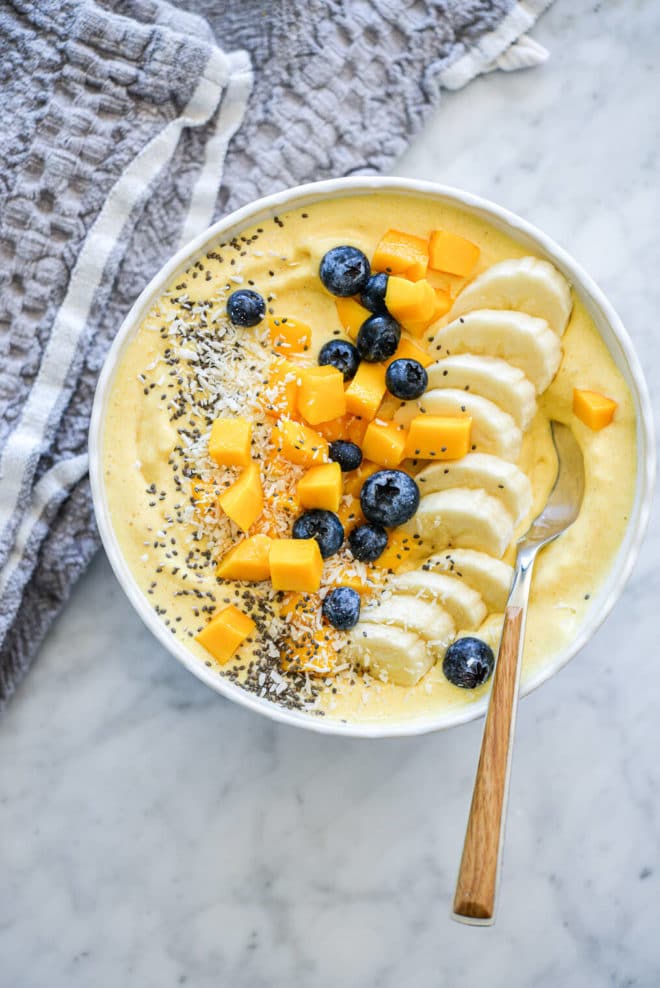 Mango smoothie with wooden and silver spoon in a bowl topped with sliced banana, chopped mango, blueberries, chia seeds, and coconut flakes