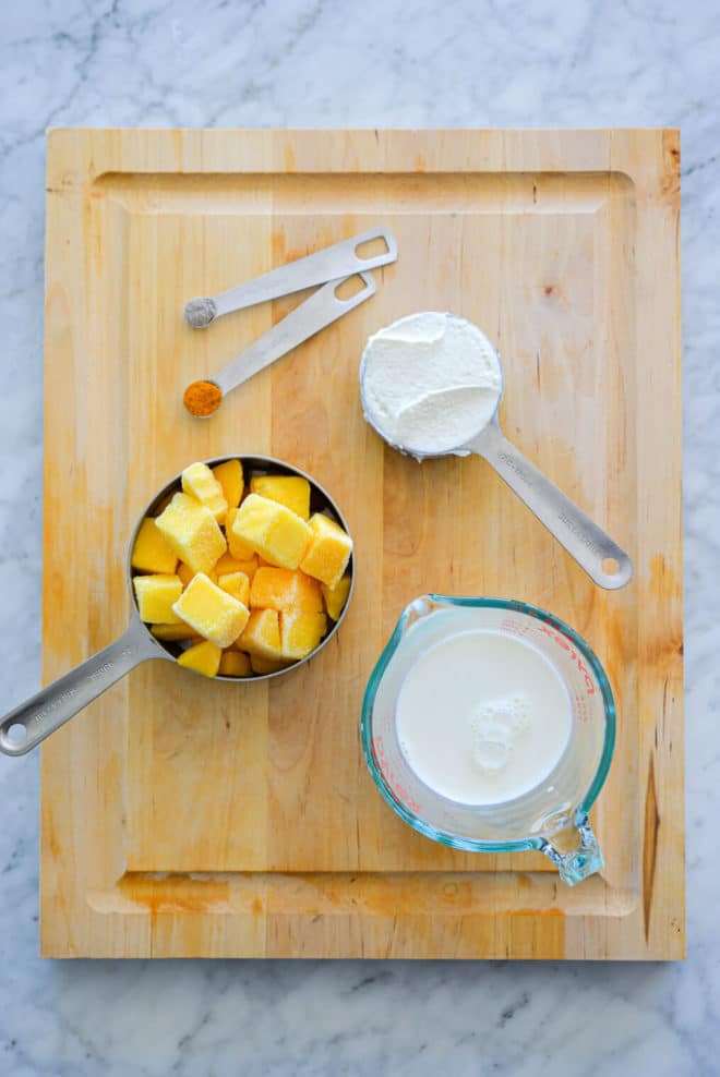 Cutting board with ground cardamon in measuring spoon, ground turmeric in measuring spoon, greek yogurt in measuring cup, frozen mango chunks in measuring cup, and milk in measuring cup