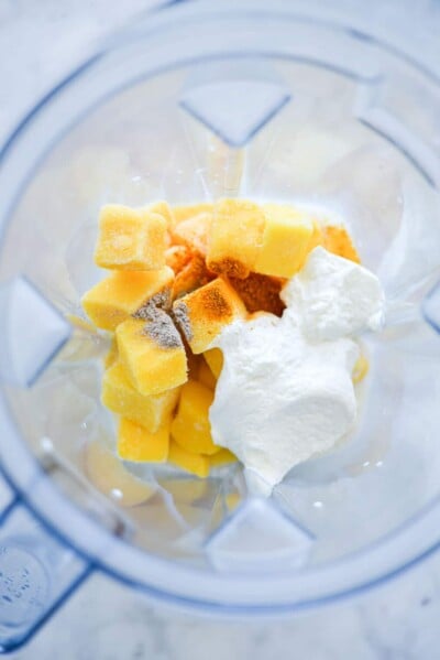 Top down view of blender with milk, frozen mango chunks, ground cardamon, ground turmeric, and greek yogurt