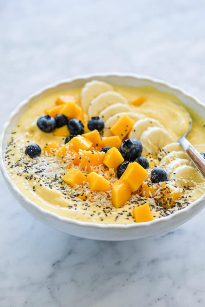 Mango smoothie with wooden and silver spoon in a bowl topped with sliced banana, chopped mango, blueberries, chia seeds, and coconut flakes