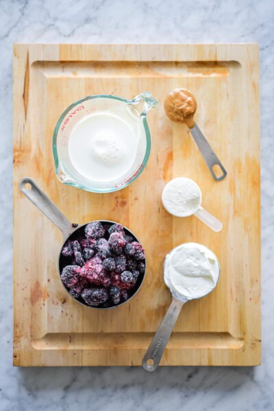 Cutting board with milk in measuring cup, frozen berries in measuring cup, peanut butter in a measuring spoon, protein powder in a measuring cup, and greek yogurt in a measuring cup