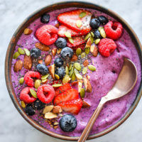 Protein smoothie with bronze spoon in bowl topped with pumpkin seeds, almonds, blueberries, raspberries, sliced strawberries, and chia seeds