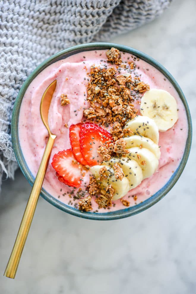 Strawberry smoothie with gold spoon in bowl topped with sliced banana, sliced strawberry, granola, and chia seeds