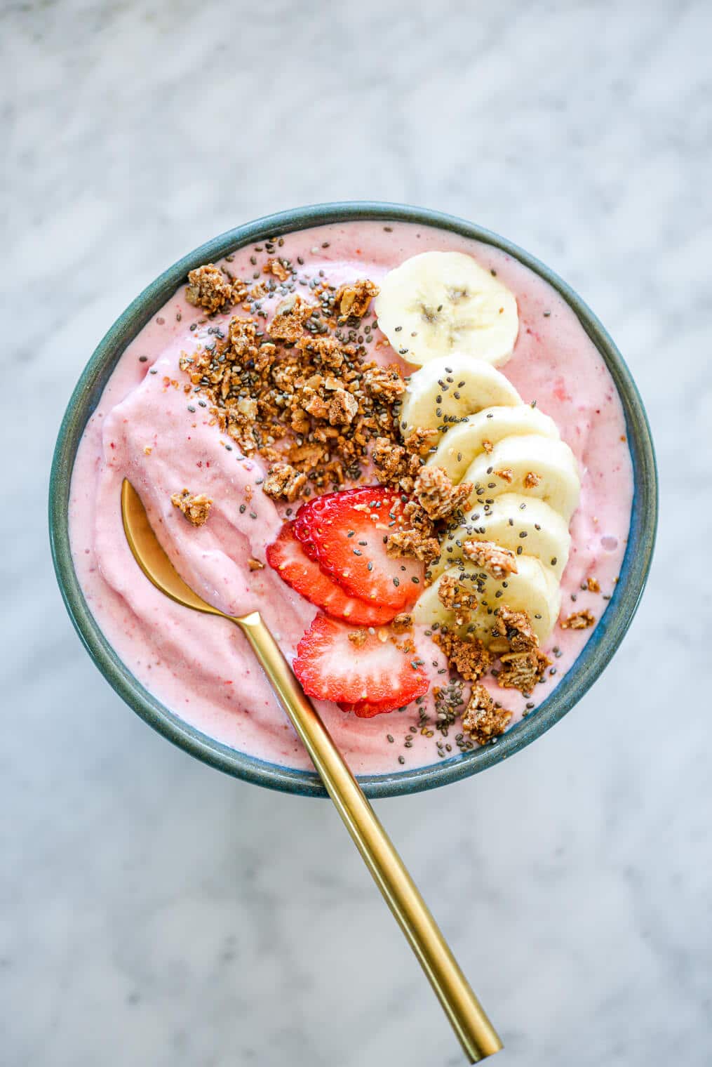 Strawberry smoothie with gold spoon in bowl topped with sliced banana, sliced strawberry, granola, and chia seeds