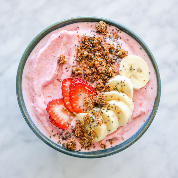 Strawberry smoothie in bowl topped with sliced banana, sliced strawberry, granola, and chia seeds