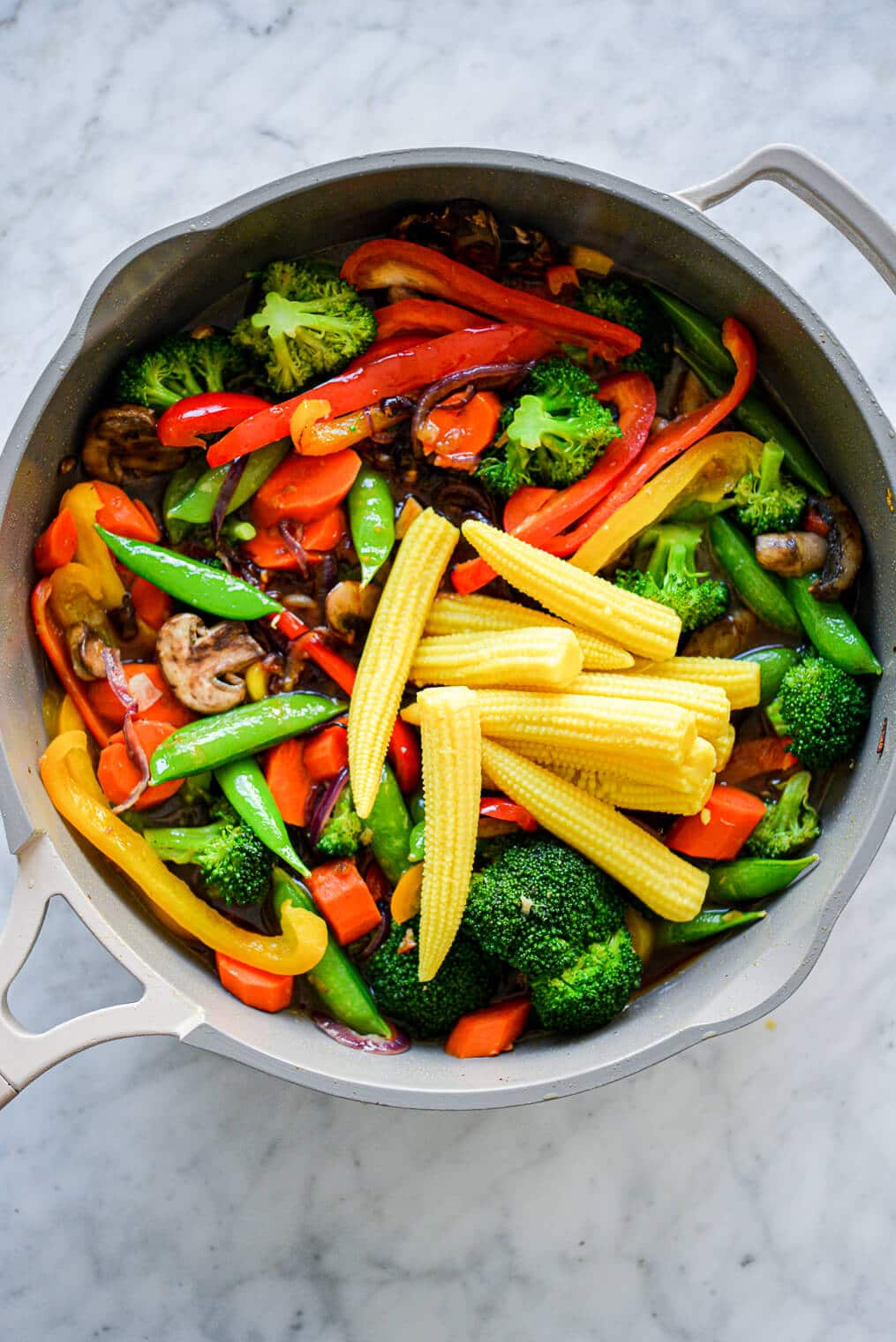 Pan with assortment of stir fry vegetables (baby corn, sugar peas, sliced red and yellow bell pepper, sliced carrots, broccoli, sliced mushrooms, and sliced red onion) sitting on grey and white marble countertop