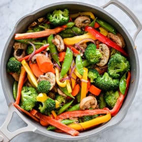 Pan with assortment of stir fry vegetables (baby corn, sugar peas, sliced red and yellow bell pepper, sliced carrots, broccoli, sliced mushrooms, and sliced red onion) sitting on grey and white marble countertop
