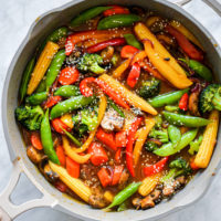 Pan with assortment of stir fry vegetables (baby corn, sugar peas, sliced red and yellow bell pepper, sliced carrots, broccoli, sliced mushrooms, and sliced red onion) sprinkled with sesame seeds sitting in a stir fry sauce