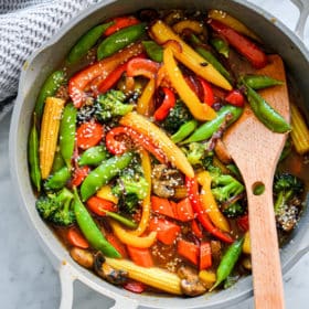 Pan with wooden spatula and assortment of stir fry vegetables (baby corn, sugar peas, sliced red and yellow bell pepper, sliced carrots, broccoli, sliced mushrooms, and sliced red onion) sprinkled with sesame seeds sitting on grey and white marble countertop with grey textured towel in the upper left corner