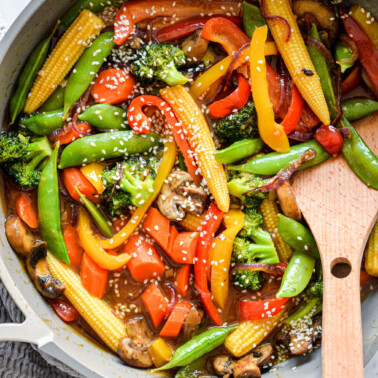 Pan with wooden spatula and assortment of stir fry vegetables (baby corn, sugar peas, sliced red and yellow bell pepper, sliced carrots, broccoli, sliced mushrooms, and sliced red onion) sprinkled with sesame seeds sitting on grey and white marble countertop with grey textured towel in the bottom left corner