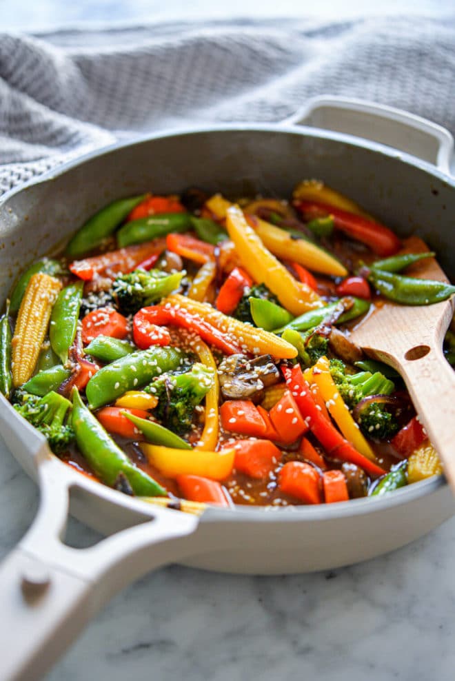 Pan with wooden spatula and assortment of stir fry vegetables (baby corn, sugar peas, sliced red and yellow bell pepper, sliced carrots, broccoli, sliced mushrooms, and sliced red onion) sprinkled with sesame seeds sitting on grey and white marble countertop with grey textured towel blurred in the upper left corner