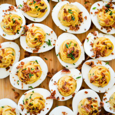 Wooden cutting board with deviled eggs topped with crumbled bacon and chives.