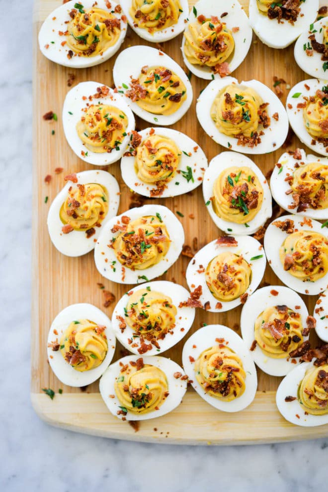 Wooden cutting board with deviled eggs topped with crumbled bacon and chives.