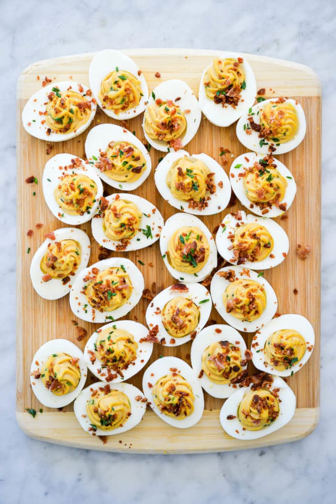 Wooden cutting board with deviled eggs topped with crumbled bacon and chives sitting on a white and grey marbled counter.