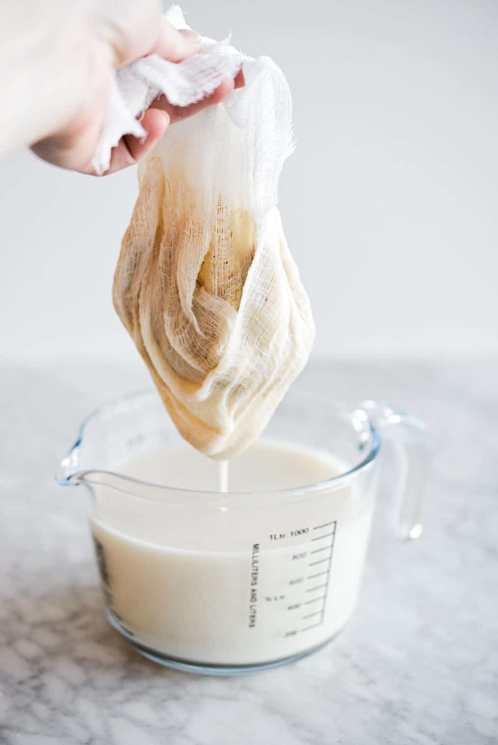 Cheese cloth with oats being held over measuring cup filled with oat milk straining from cheese cloth.