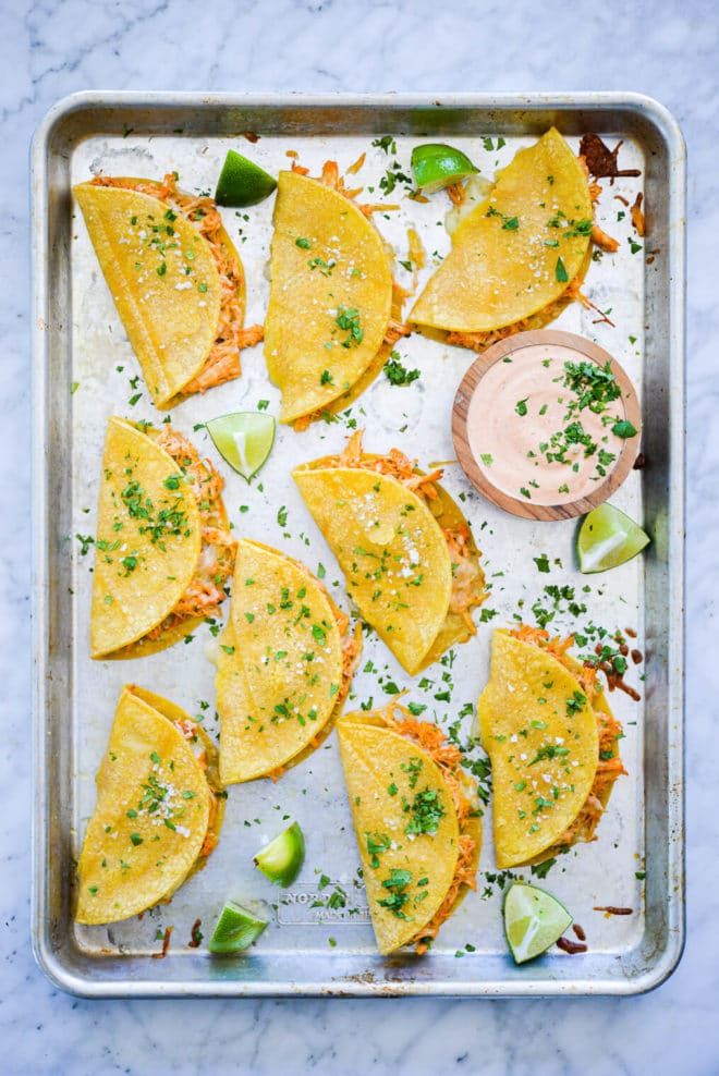 Sheet pan with cooked shredded chicken tacos topped with chopped cilantro, quarters of fresh lime, and a small, wooden bowl with creamy dipping sauce.