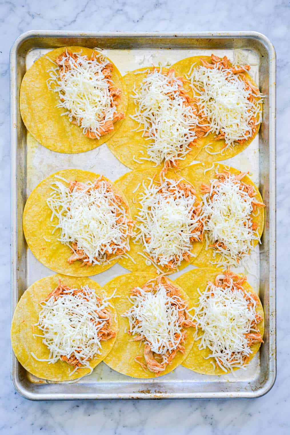 Sheet pan with three rows of three corn tortillas. Each tortilla is topped on one half side with shredded chicken and shredded cheese.