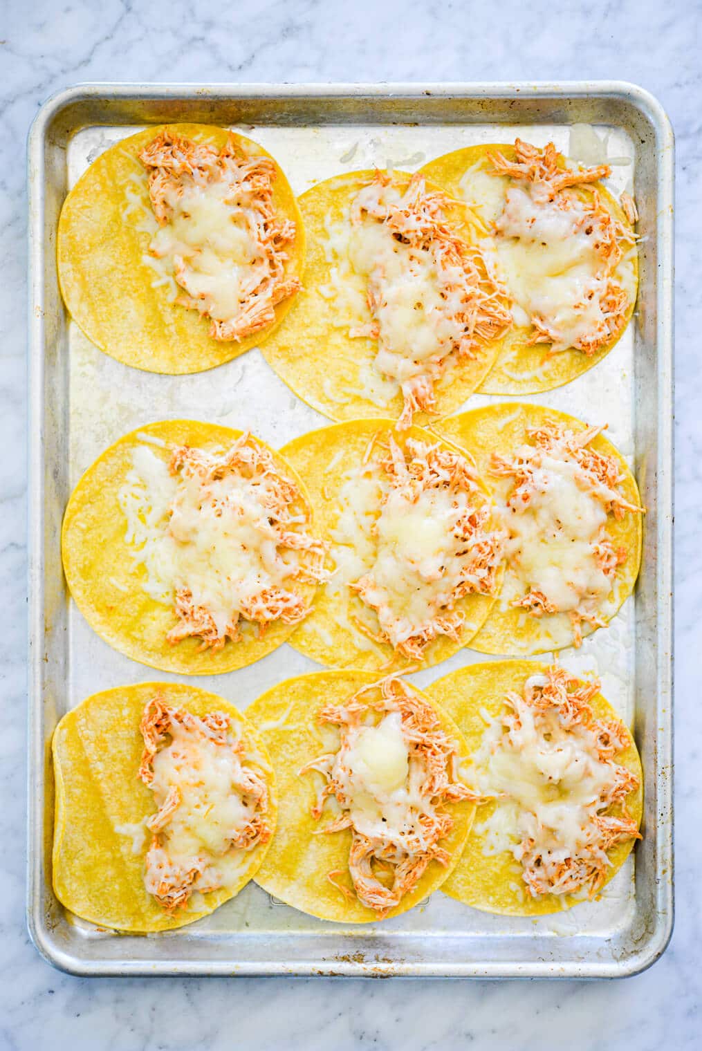 Sheet pan with three rows of three corn tortillas. Each tortilla is topped on one half side with shredded chicken and melted shredded cheese.