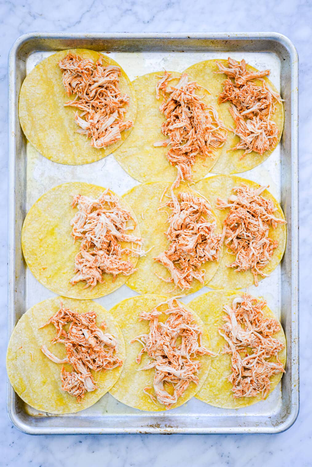 Sheet pan with three rows of three corn tortillas. Each tortilla is topped on one half side with shredded chicken.