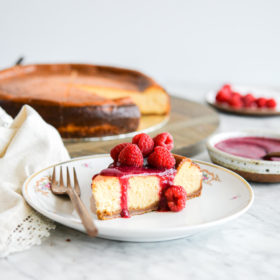 Slice of classic cheesecake topped with raspberry jam and whole raspberries sitting on a gold-rimmed, white plate with small magenta flowers in floral design and a copper fork with full cheesecake and plate of raspberries blurred in the background.