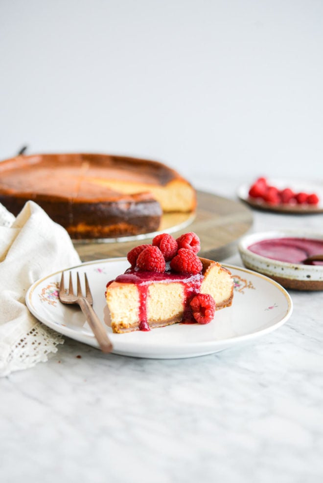 Slice of classic cheesecake topped with raspberry jam and whole raspberries sitting on a gold-rimmed, white plate with small magenta flowers in floral design and a copper fork with full cheesecake and plate of raspberries blurred in the background.