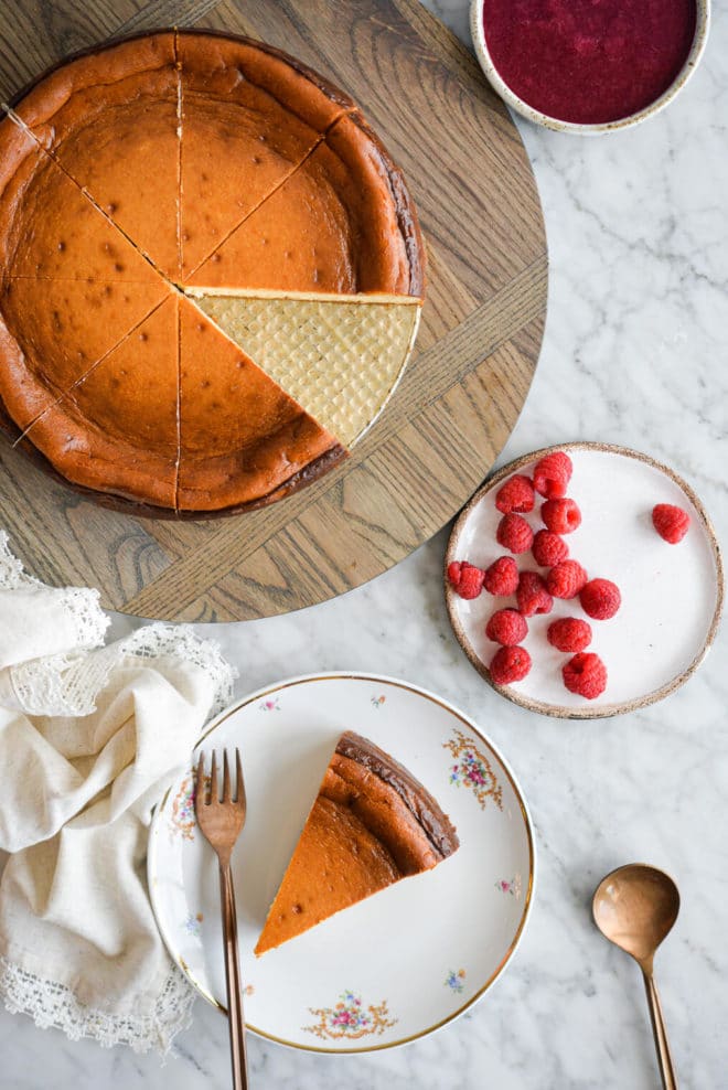 Top down view of slice of classic cheesecake sitting on a gold-rimmed, white plate with small magenta flowers in floral design and a copper fork with full cheesecake, plate of raspberries, and bowl of raspberry jam.
