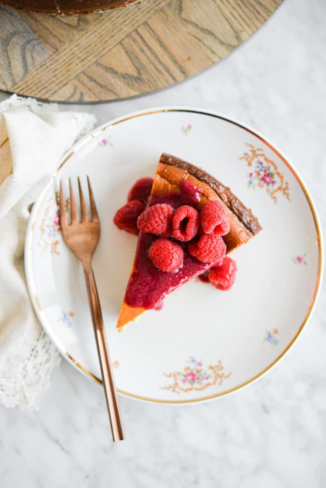 Slice of classic cheesecake topped with raspberry jam and whole raspberries sitting on a gold-rimmed, white plate with small magenta flowers in floral design and a copper fork.