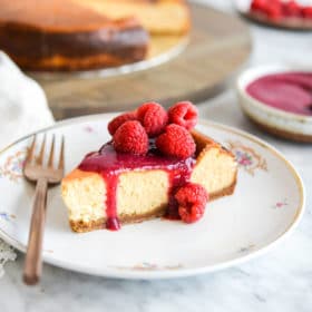 Slice of classic cheesecake topped with raspberry jam and whole raspberries sitting on a gold-rimmed, white plate with small magenta flowers in floral design and a copper fork with full cheesecake and plate of raspberries blurred in the background.