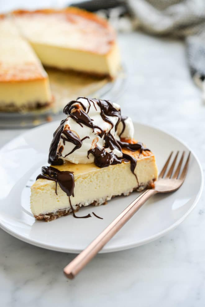 Slice of cheesecake with whipped cream and melted chocolate drizzled on top on a white plate with copper fork and full cheesecake blurred in the background.