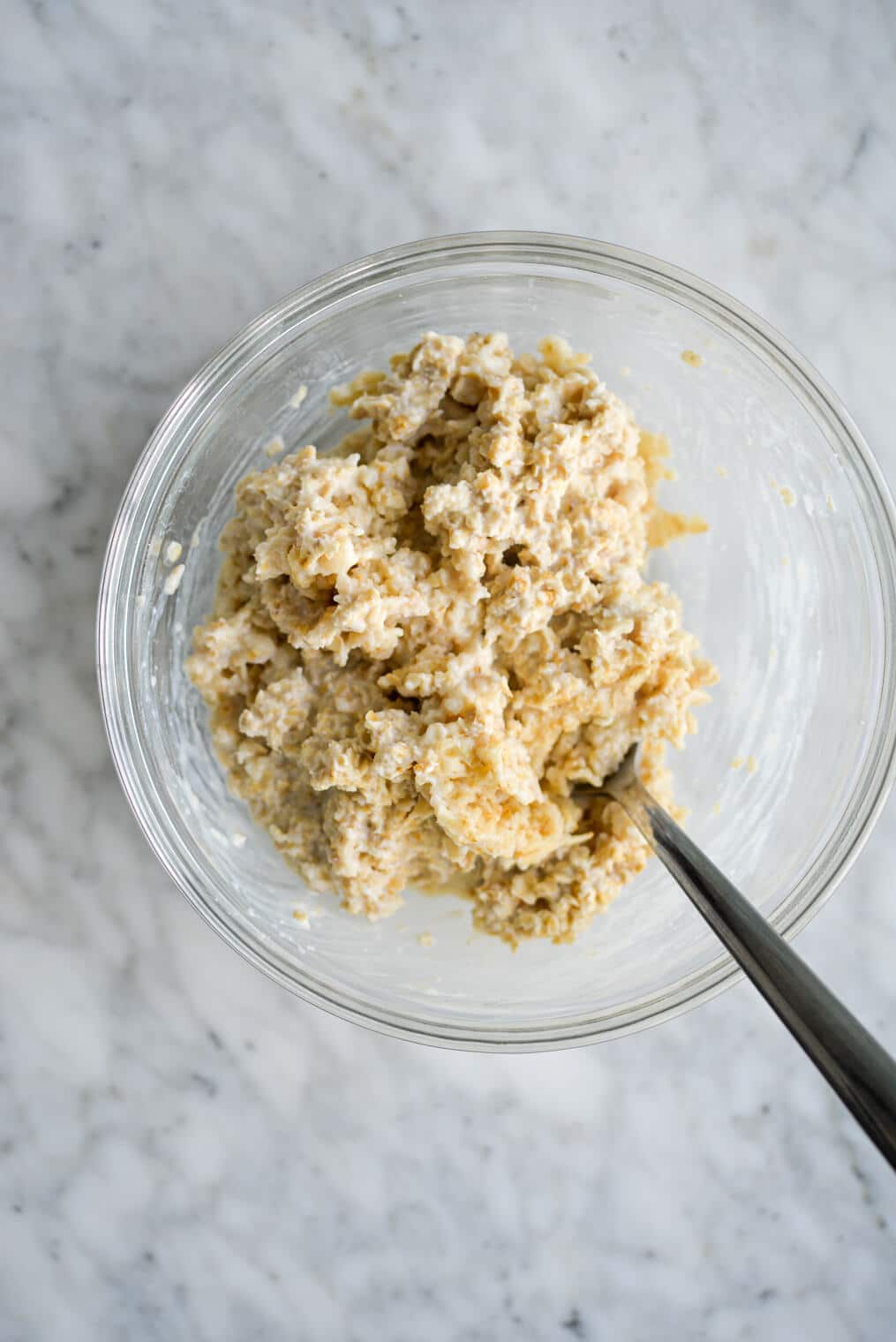 Heated oatmeal stirred with a metal spoon in a glass bow sitting on a marble countertop.