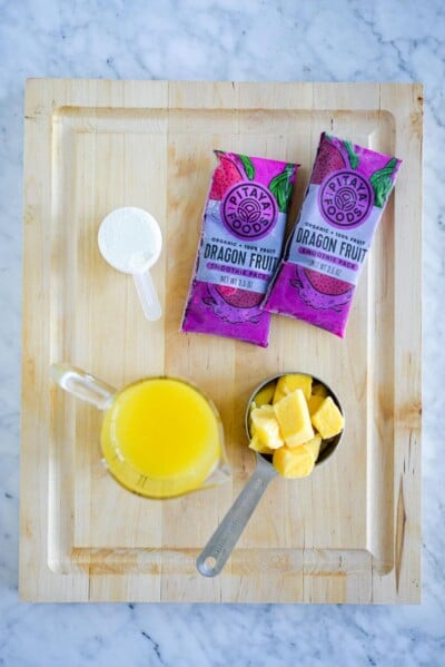 Ingredients for pitaya smoothie bowl sitting on a rectangle, wooden cutting board.