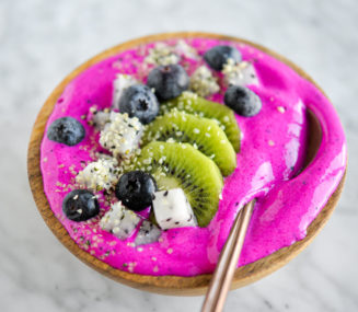 Bright magenta smoothie topped with sliced kiwi, dragonfruit, and blueberries in a wooden bowl with a spoon.