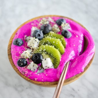 Bright magenta smoothie topped with sliced kiwi, dragonfruit, and blueberries in a wooden bowl with a spoon.