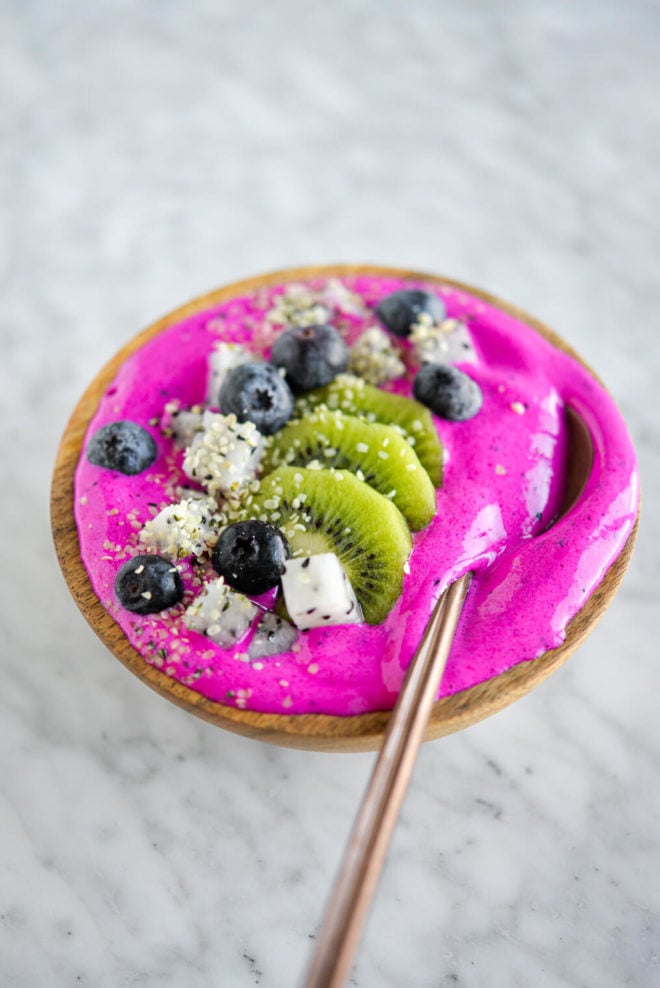 Bright magenta smoothie topped with sliced kiwi, dragonfruit, and blueberries in a wooden bowl with a spoon.