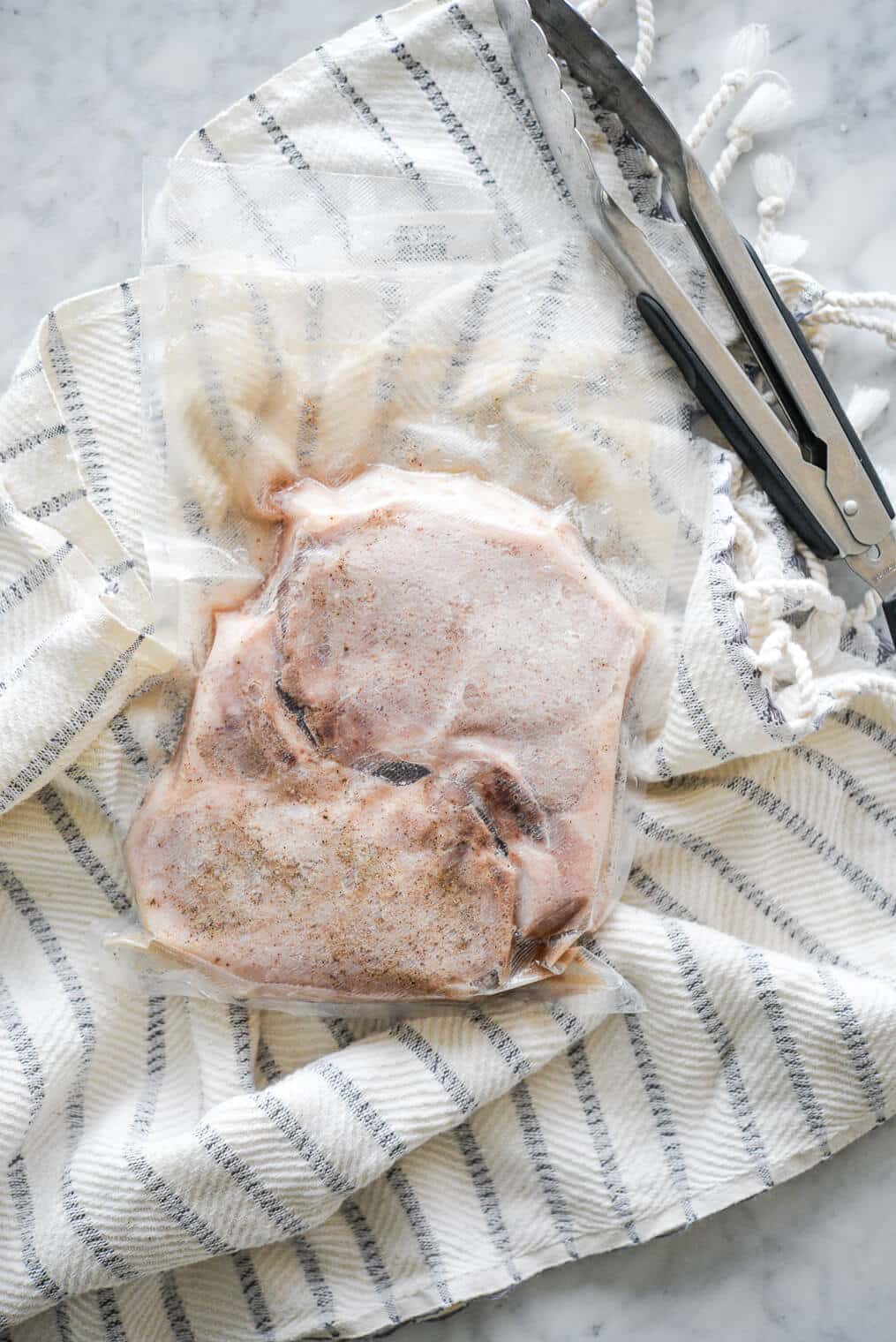 Two cooked, bone-in pork chops in a sous vide bag sitting next to a pair of tongs on top of a white and blue striped towel.