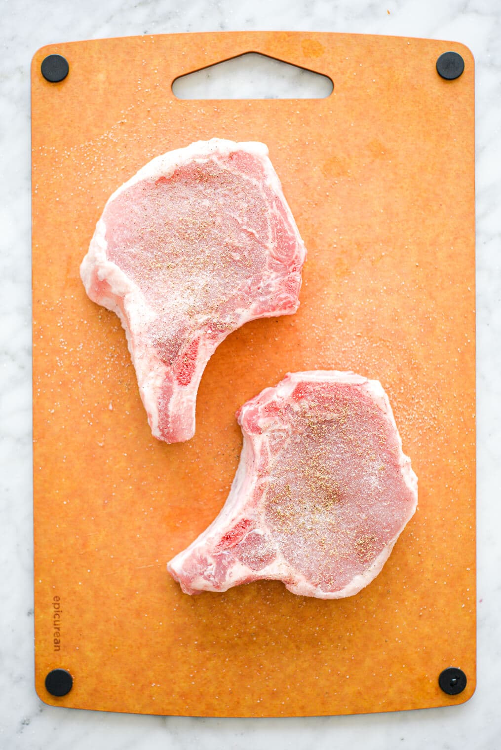 Two, raw bone-in pork chops on a cutting board seasoned with salt and pepper.