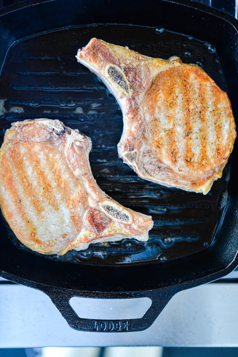 Two bone-in pork chops searing in a square cast iron grill pan.