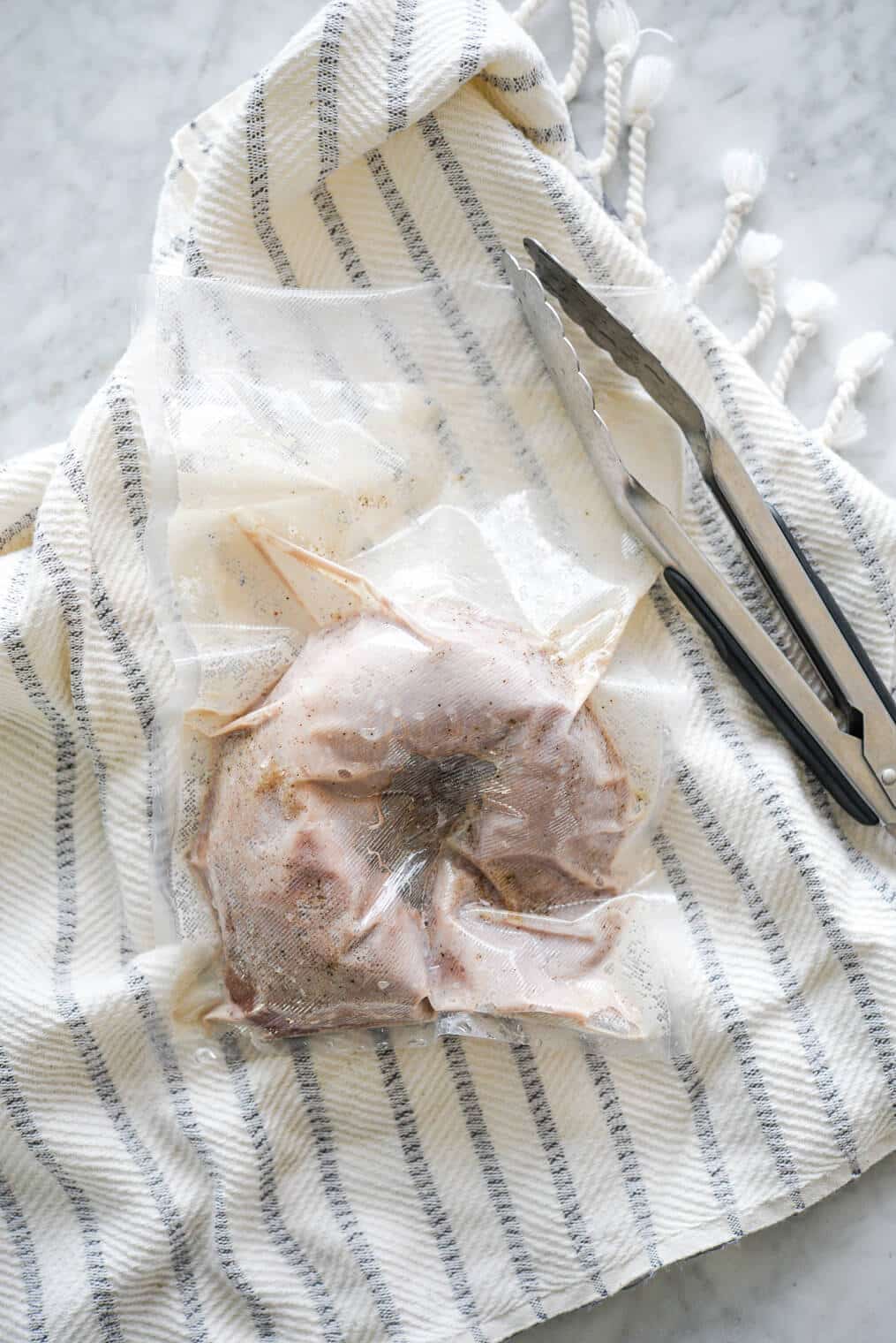 Pork tenderloin in a vacuum sealed bag sitting next to a pair of tongs on top of a white and blue striped towel.