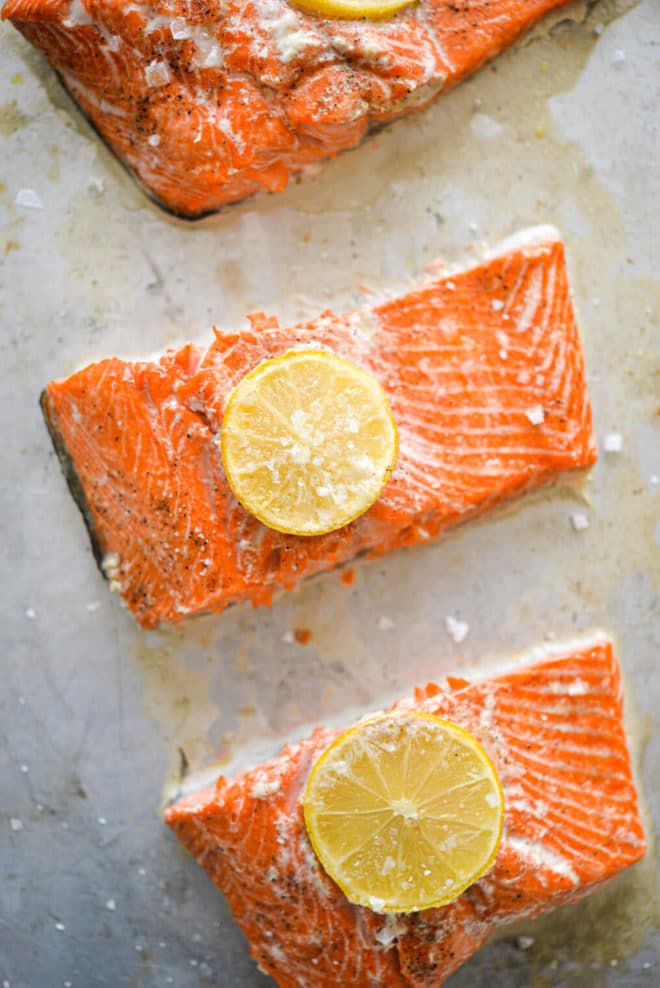 Three sliced salmon filets topped with lemon slices on a metal baking sheet.