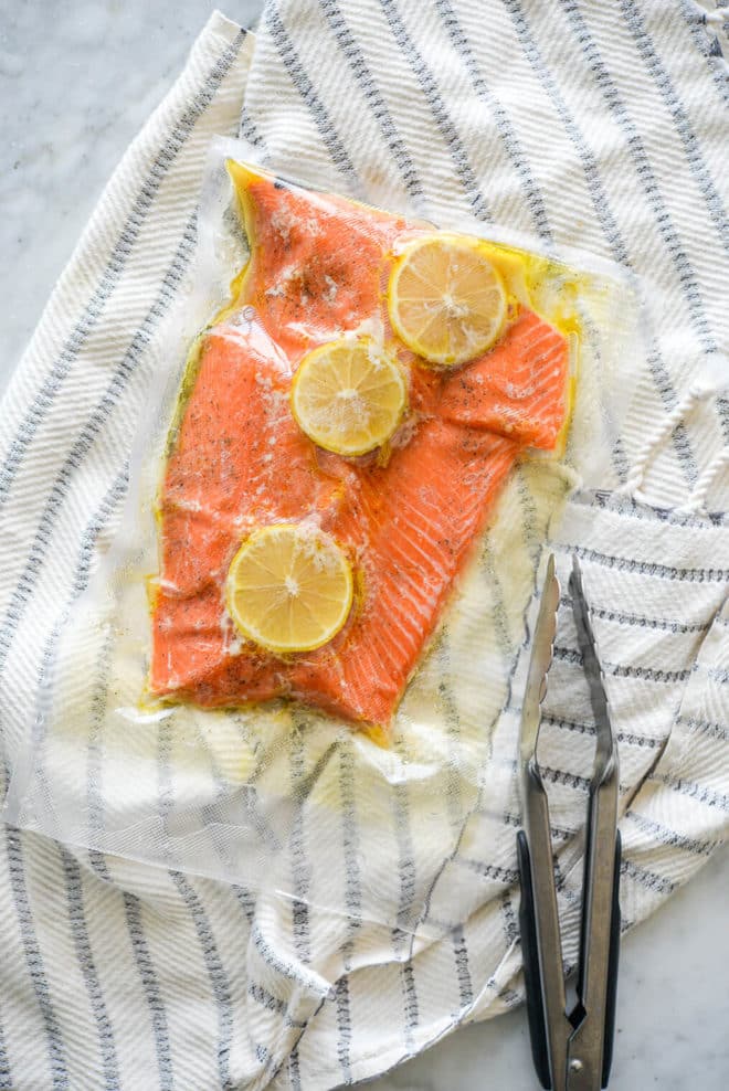 Vacuum sealed bag with salmon filet topped with lemon slices on top of a blue and white striped towel with a pair of tongs to the bottom right.