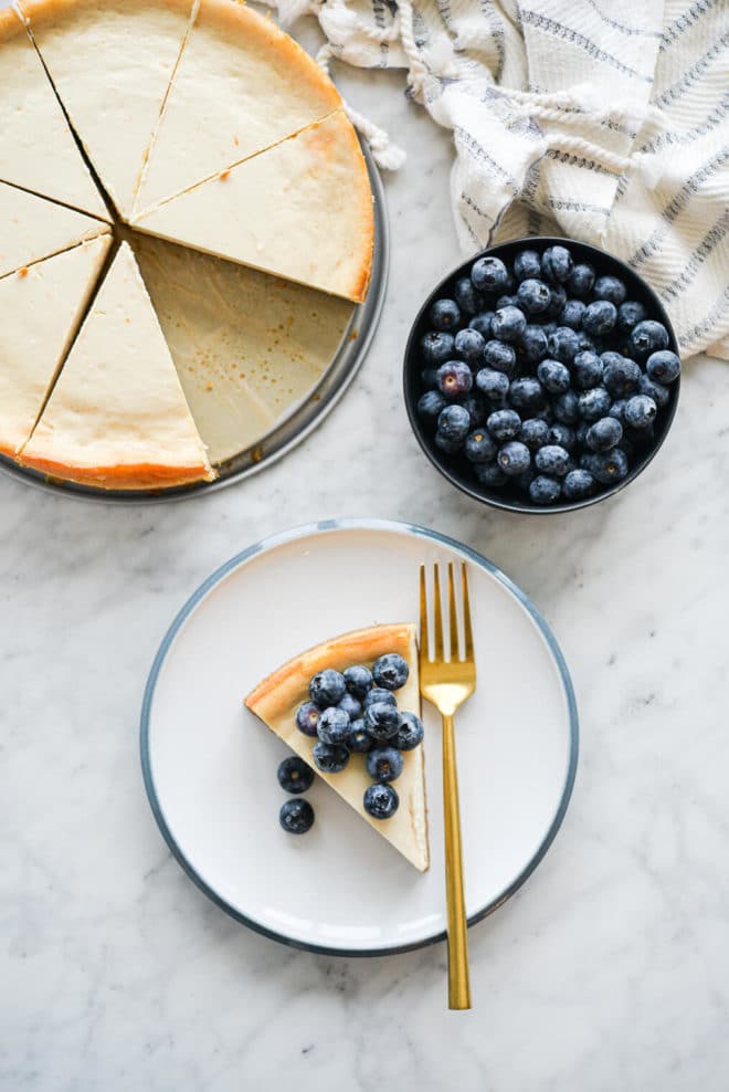 Slice of cheesecake topped with blueberries on a white plate with blue rim with whole cheesecake and bowl of blueberries.