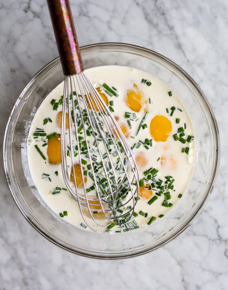 Glass bowl with eggs, cream, chives, and a whisk.