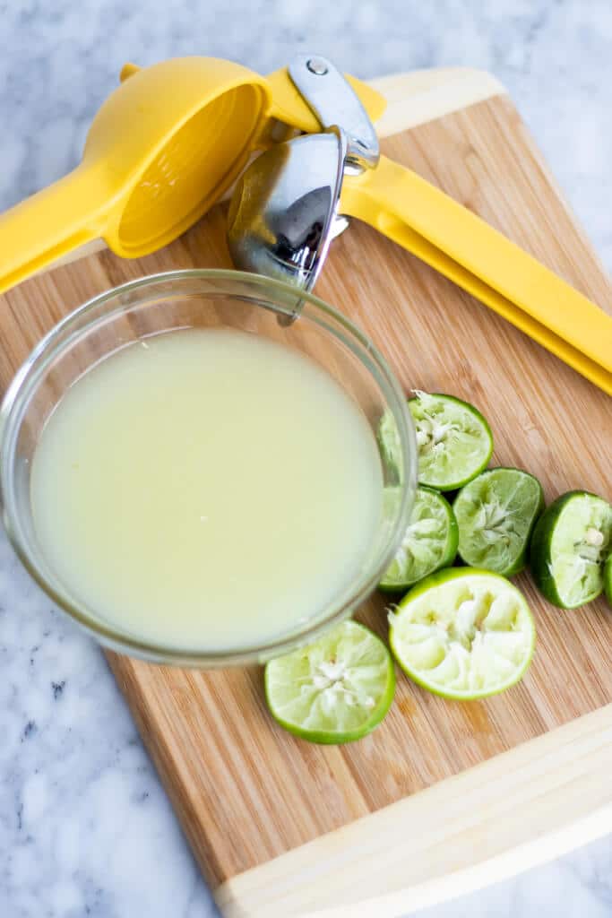 Citrus juicer, glass bowl with lime juice, and juiced key lime halves on a wooden cutting board.
