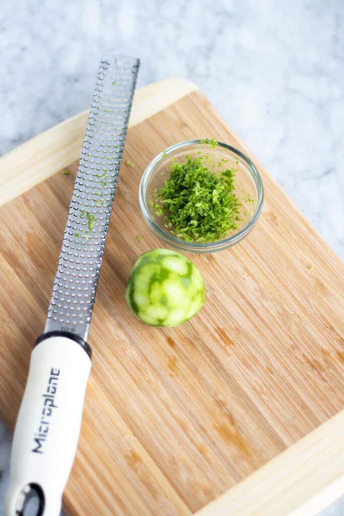 Microplane with key lime and small glass bowl with lime zest on a wooden cutting board.