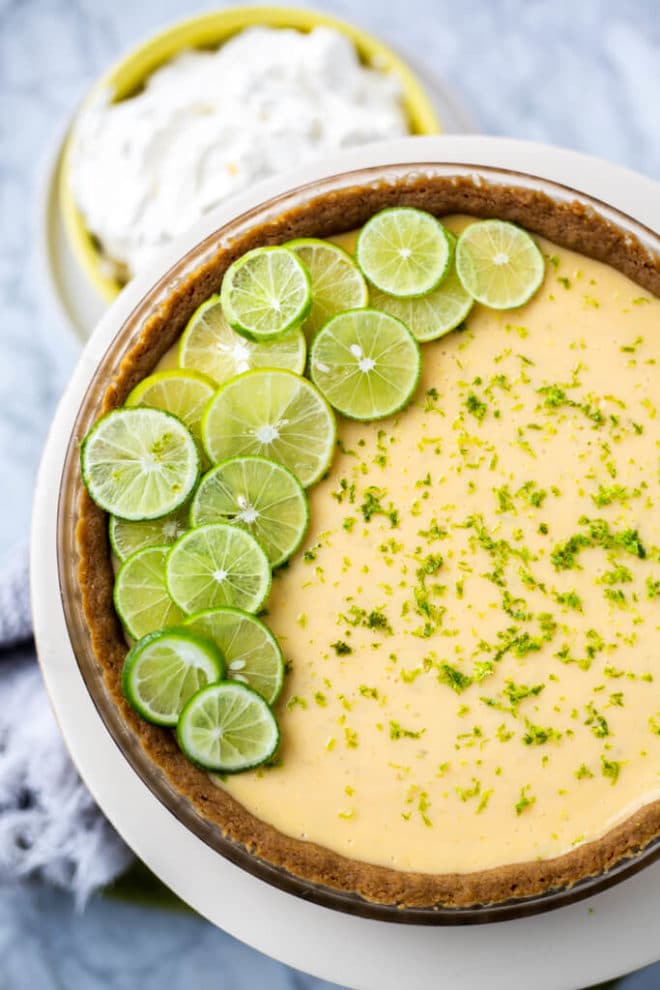 Top down view of baked key lime pie topped with lime zest and sliced key limes layered on one side sitting on a white cake stand with a bowl of whipped cream below.