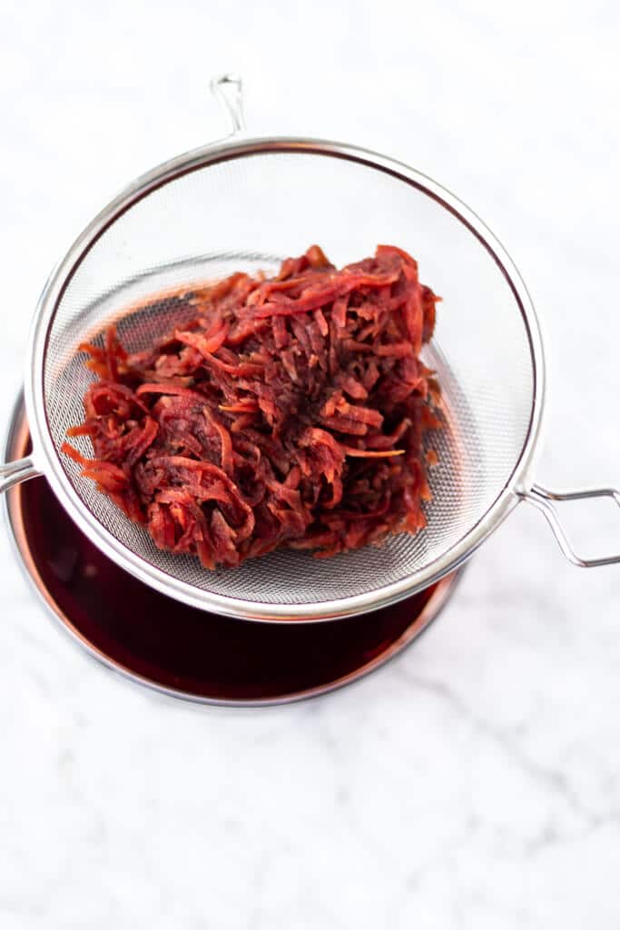 Shredded beets in a metal sieve held over a glass jar filled with liquid.