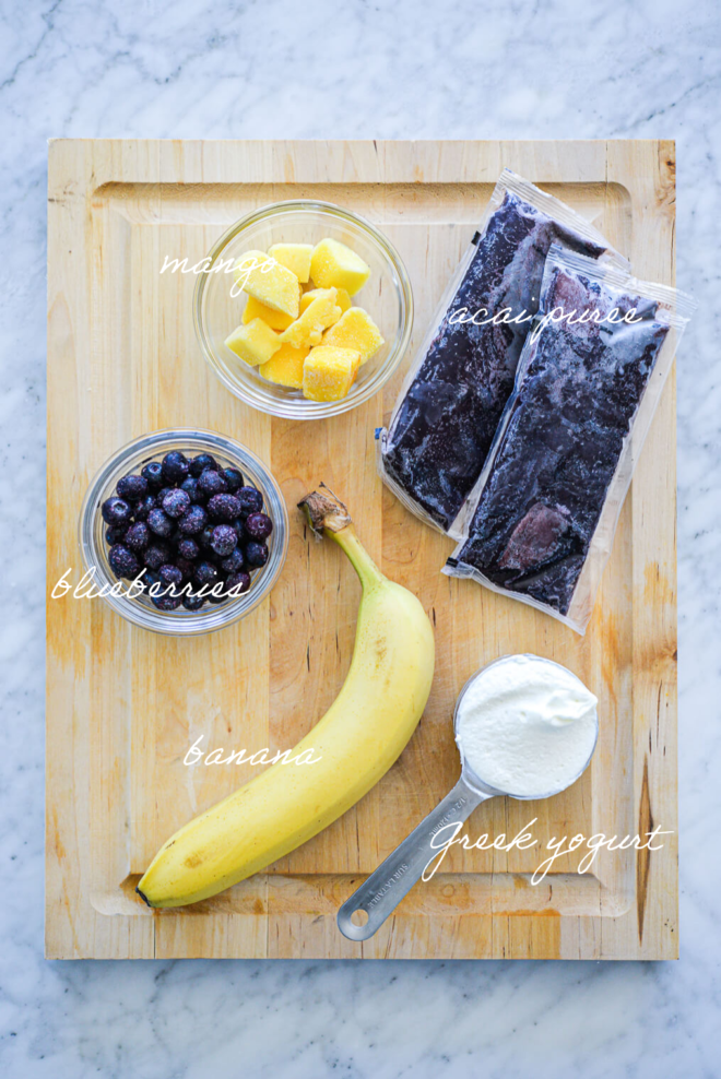 Acai smoothie ingredients on a wooden cutting board with ingredient labels written in white letters.